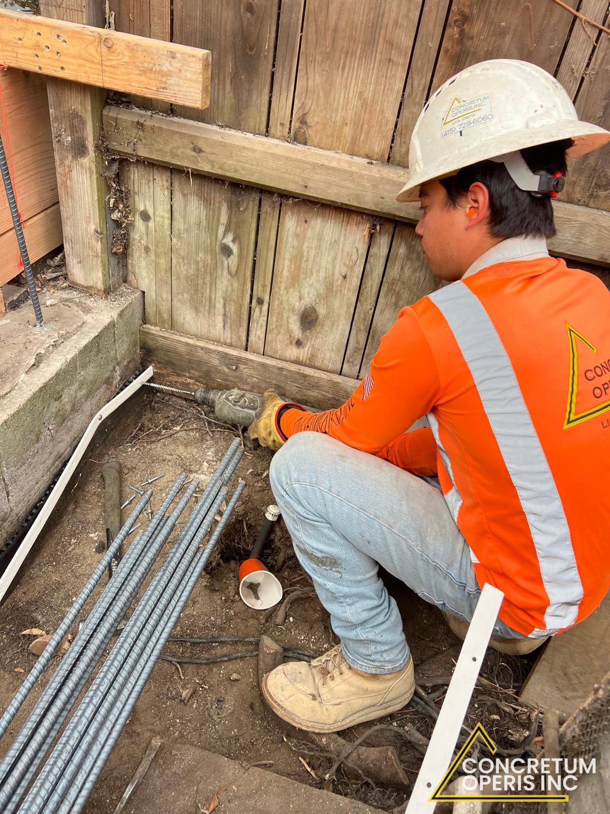 Concretum Operis crew member drilling rebar dowels into a footing near a wood fence