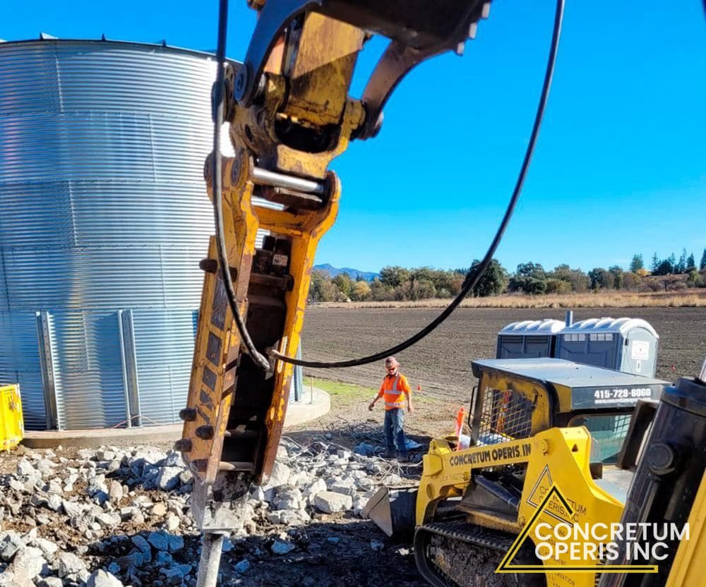 Excavator-mounted hydraulic hammer breaking concrete rubble near a water tank in Richmond, CA